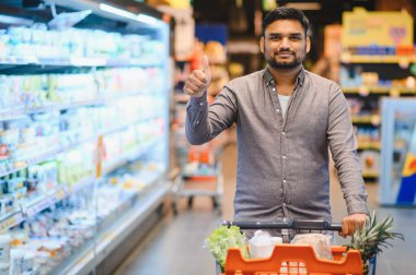 Mutlu adam dolu bir alışveriş arabasını itiyor, süpermarket deneyiminden tatmin olduğunu ifade ediyor, pozitif müşteri seçimi gösteriyor.