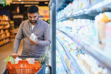 Genç adam süt şişesinde etiket okuyor, marketleri buzdolabı koridorunda erzakla dolduruyor.