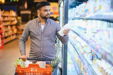 Erkek müşteri alışveriş arabasını koridor boyunca itiyor, süt şişesini tutuyor ve şeffaf buzdolabında etiketini kontrol ediyor.
