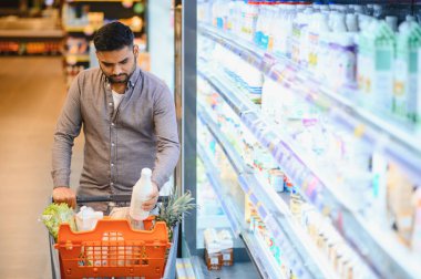 Genç adam alışveriş arabasını itiyor, buzdolabından süt şişesi alıyor, markette sağlıklı yiyecek seçimleri yapıyor.