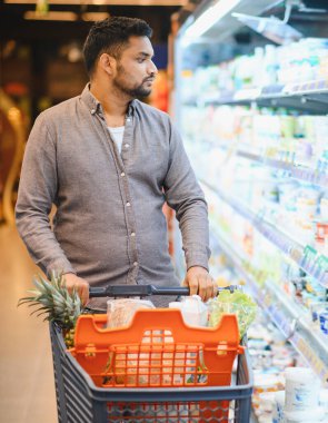 Taze ürünler de dahil olmak üzere bir alışveriş arabasını iten adam, bir süpermarket koridorunda ürünleri tarıyor.