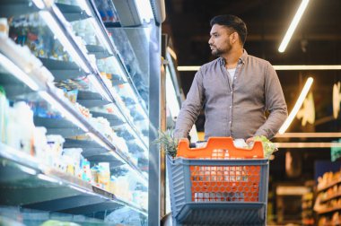 Alışveriş arabalarını soğutuculu süpermarket koridorlarında iten adam günlük market alışverişi sırasında yiyecek ve içecek seçiyor.