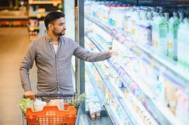 Genç adam alışveriş arabasını itiyor, marketin soğutulmuş mandırasında süt veya yoğurda uzanıyor.