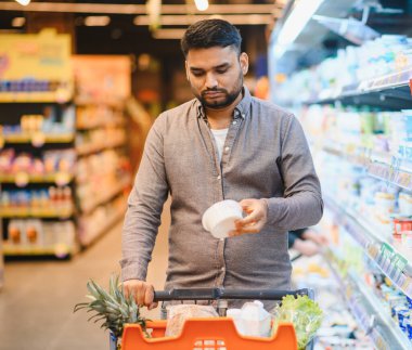 Süpermarketteki genç adam bir ürün etiketini inceliyor, beslenme ve fiyatı karşılaştırırken düşünceli bir market seçimi yapıyor.