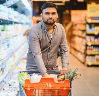 Genç adam canlı bir süpermarket koridorunda taze ürünlerle dolu bir alışveriş arabasını itiyor.