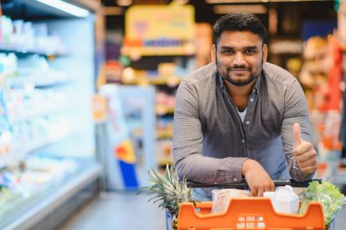 Mutlu adam gülümsüyor, markette içi taze ürünlerle dolu bir alışveriş arabasını itiyor, alışveriş deneyiminin tadını çıkarıyor.