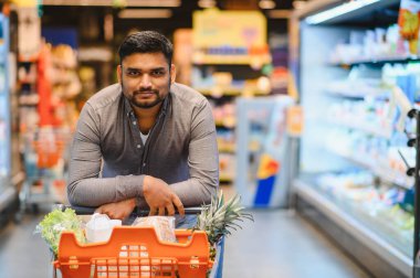 Süpermarket koridorunda içi taze ürünlerle dolu bir alışveriş arabasıyla poz veren adam sağlıklı bir yaşam tarzına hazır.