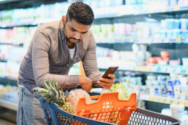 Akıllı telefondan alışveriş listesini kontrol eden bir adam market koridorunda alışveriş arabasının üzerine yaslanıyor. Akıllı ticaret kavramı