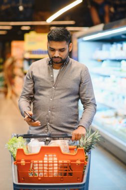 Erzak dolu alışveriş arabasını iten adam, akıllı telefonuyla modern bir marketteki ürünleri doğrulamak için başvuru yapıyor.