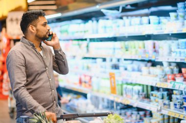 Alışveriş arabasını iten bir adam, manav koridorundaki bir cep telefonuyla süt ürünleriyle iletişim kuran bir adam.