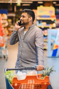 Süpermarkette alışveriş yapan genç adam, cep telefonuyla konuşurken gülümseyen market alışverişi yapan, perakende koridorunda gezinen.
