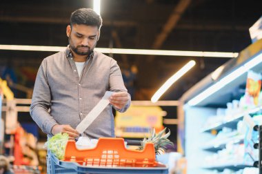 Genç adam yiyecek alışverişi yaptıktan sonra uzun bir market fişini inceliyor ve parlak ışıklandırılmış bir süpermarkette üretim yapıyor.