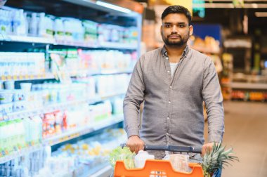 Taze ürünlerle dolu bir alışveriş arabasını iten adam, parlak ışıklandırılmış bir markette ürünleri karıştırıyor.