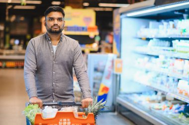 Genç adam bakkalda alışveriş yapıyor, içi taze ürün ve yiyecek dolu bir süpermarket arabasını itiyor.