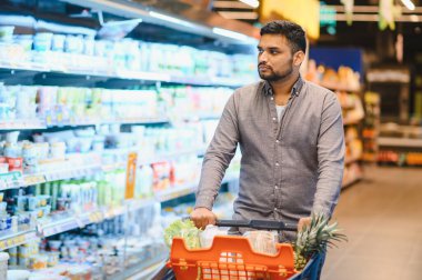 Süpermarket koridorlarında alışveriş arabası süren, yiyecek seçen bir adam. Taze yiyecek ve süt ürünleri alan tüketici