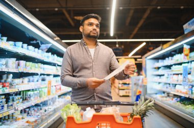 Genç adam uzun bir alışveriş listesini dikkatlice incelerken bir yandan da alışveriş arabasının açık bir süpermarket koridorunda market ihtiyaçlarına odaklanıyor.