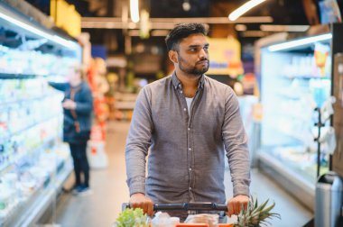 Genç adam market alışverişi yapıyor, içi taze ürünlerle dolu bir arabayı parlak bir süpermarket koridorunda itiyor.