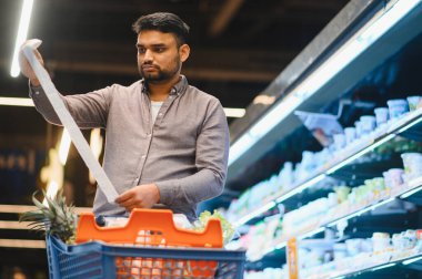 Hintli bir adam, uzun bir alışveriş fişini inceliyor. Parlak bir market koridorunda, yemek arabasıyla harcamaları değerlendiriyor.