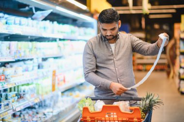 Market koridorunda duran genç adam, bir el arabasını iterken uzun bir kağıt makbuzunu ya da alışveriş listesini dikkatlice inceliyor.