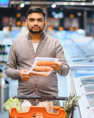 Süpermarketteki genç adam taze ürünlerle dolu bir arabayı iterken paketlenmiş balığı inceliyor ve düşünceli bir satın alma kararı veriyor.