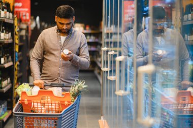Market koridorundaki bir ürün paketinin malzemelerini ya da son kullanma tarihlerini karşılaştırmak için alışveriş arabasını iten adam.