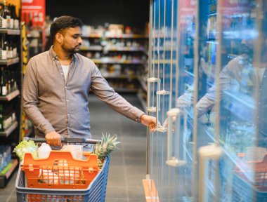 Alışveriş arabalı bir adam soğutuculu vitrinden eşyalar seçiyor, alışveriş deneyimiyle meşgul oluyor.