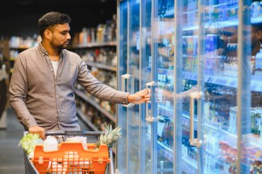 Genç adam markette alışveriş yapıyor, alışveriş arabası itiyor ve dondurulmuş bir kapı açıyor.