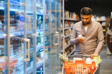 Müşteri buzdolabı rafından bir ürün seçiyor, içi taze yiyecek ve temel malzemelerle dolu bir alışveriş arabasını itiyor.