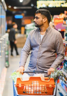 Genç bir adam, perakende mağazası koridorunda ürünleri seçerken içi taze ürünlerle dolu bir alışveriş arabasını itiyor.