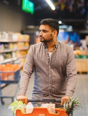 Genç adam, modern bir markette içi taze ürünlerle dolu bir alışveriş arabasını itiyor.