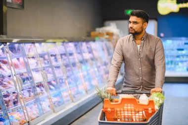 Bir marketteki soğutuculu vitrinler boyunca içi taze ürün ve süt ürünleriyle dolu bir alışveriş arabasını iten adam.