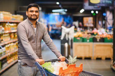 Alışveriş yaparken gülümseyen bir adam, modern bir süpermarkette parlak bir koridorda dolu bir arabayı iterken.