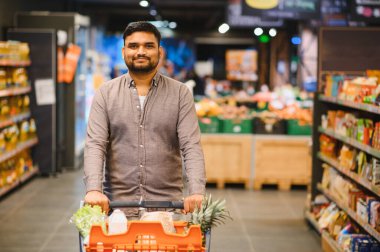 Genç adam, alışveriş arabasını iterken gülümsüyor ve modern bir süpermarket koridorunda alışveriş yapıyor.