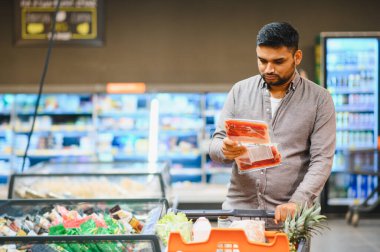 Satılık taze eti elinde tutan bir adam süpermarket koridorlarında alışveriş arabasıyla seçenekleri karşılaştırıyor.