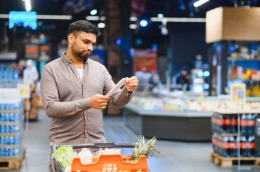 Adam alışveriş listesini süpermarkette yürürken alışveriş arabasındaki malzemelerle karşılaştırıyor, bütçe ve seçime odaklanıyor.