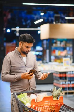 Modern bir süpermarket koridorunda market arabasını iterken akıllı telefondan dijital ve kağıt alışveriş listesini kontrol eden bir adam.