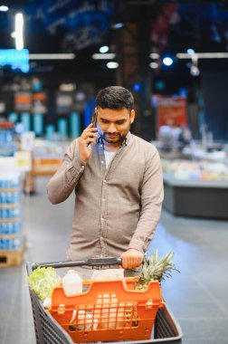 Markette cep telefonuyla konuşurken yiyecek dolu market arabasını iten genç bir adam.