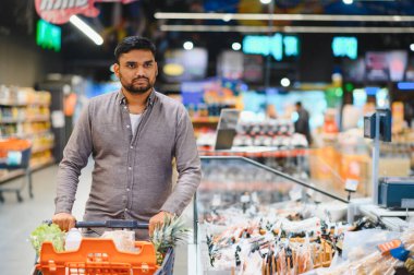 Turuncu bir alışveriş arabasını iten Hintli bir adam alışveriş yaparken ileriye bakan geniş bir süpermarket koridorunda taze ürünlerle dolu.