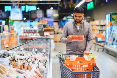Elinde paketlenmiş et ürünü olan bir adam, alışveriş arabasıyla parlak bir market koridorunda yürüyor, satın alma kararı veriyor.