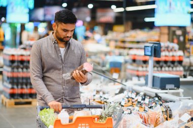 Market alışverişi yapan, elinde bir paket sosis tutan ve incelerken alışveriş arabasını iten bir adam.