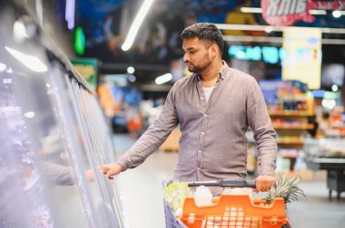 Süpermarketin dondurucu koridorunda dondurulmuş yiyeceklere bakarken alışveriş arabalarını iten adam, market gezisi için ürünleri seçiyor.