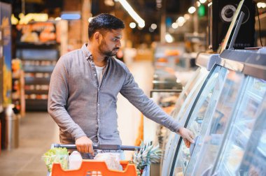 Genç adam bir alışveriş arabasını itiyor ve modern bir markette soğutuculu bir vitrinden eşyalar seçiyor.