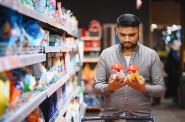 Süpermarkette dikilen, elinde iki şişe sprey tutan ve farklı temizlik ürünlerini karşılaştıran bir adam. Tüketici seçimi ve alışveriş kavramı