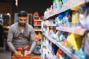 Elinde sprey şişeleri olan ve marketteki bir raftan temizlik ürünleri seçen bir adam alışveriş arabasını dolduruyor.
