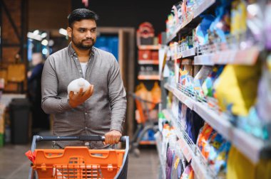 Alışveriş arabasını iten ve raflardaki temizlik ürünlerini karşılaştıran bir adam bir markette satın alma kararı veriyor.