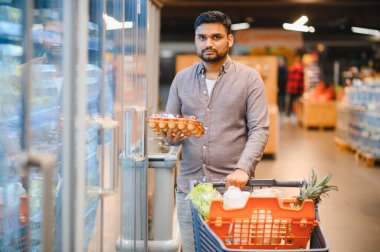 Genç Hintli adam süpermarket koridorunda alışveriş yapıyor, elinde bir karton yumurta tutuyor ve odaklanmış bir şekilde alışveriş arabasını itiyor.
