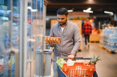 Süpermarketin soğutulmuş bölümünden taze yumurta alan adam. Alışveriş arabası market malzemeleriyle dolu.