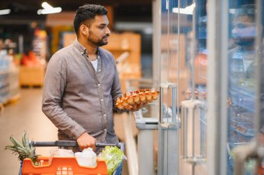 Genç Hintli bir adam, modern bir süpermarket koridorunda alışveriş arabasını iterken ürünleri ve fiyatları karşılaştırırken düşünceli bir şekilde erzak seçiyor.