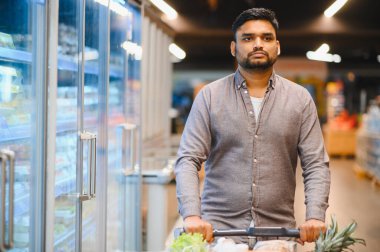 Genç bir adam, modern bir süpermarkette dondurulmuş gıda reyonlarında günlük market alışverişine odaklanmış bir el arabasını itiyor.