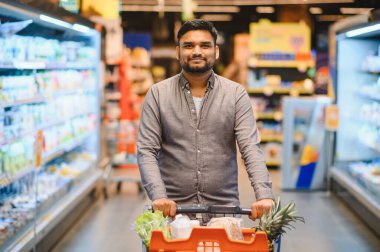 Taze ürünler ve paketlenmiş ürünlerle dolu bir alışveriş arabasını kalabalık bir market koridorunda iten adam.